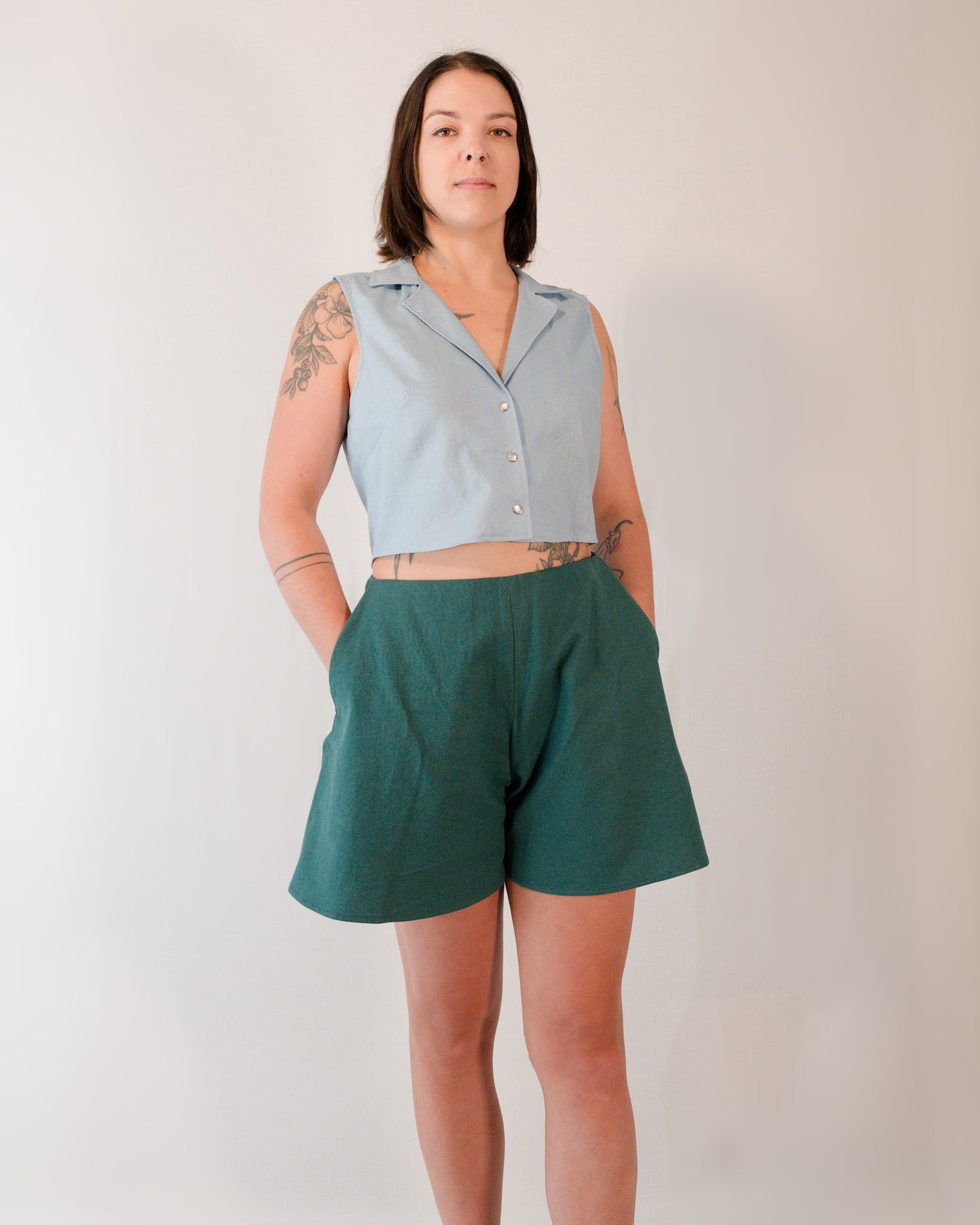 Flared shorts in deep emerald. Model has one hand in each pocket. Paired with a sleeveless blouse in skyline blue.