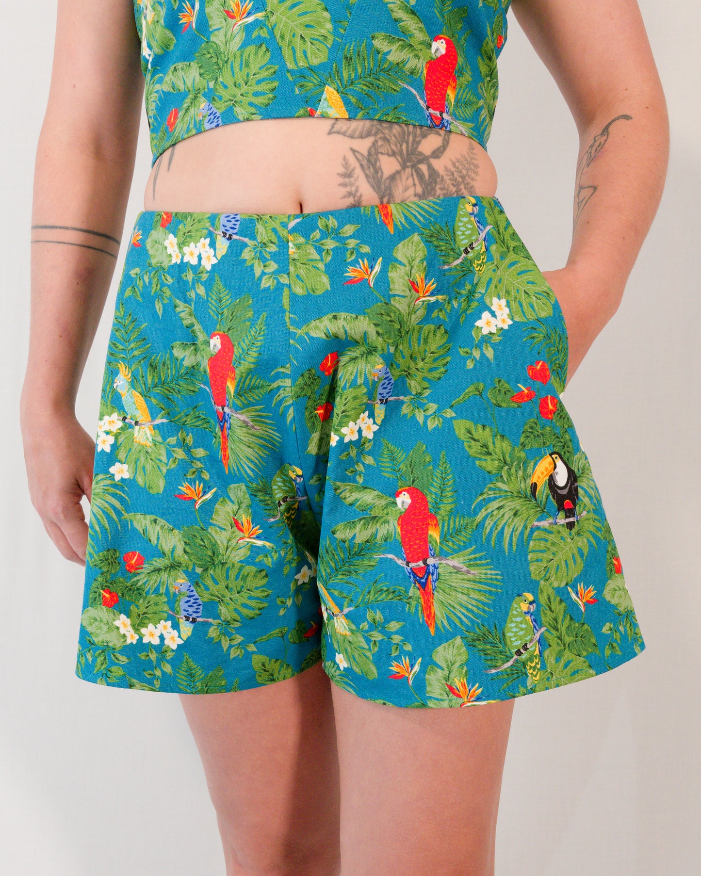 Front of flared shorts in a turquoise print with plants, parrots and toucans. Model has hand in pocket.