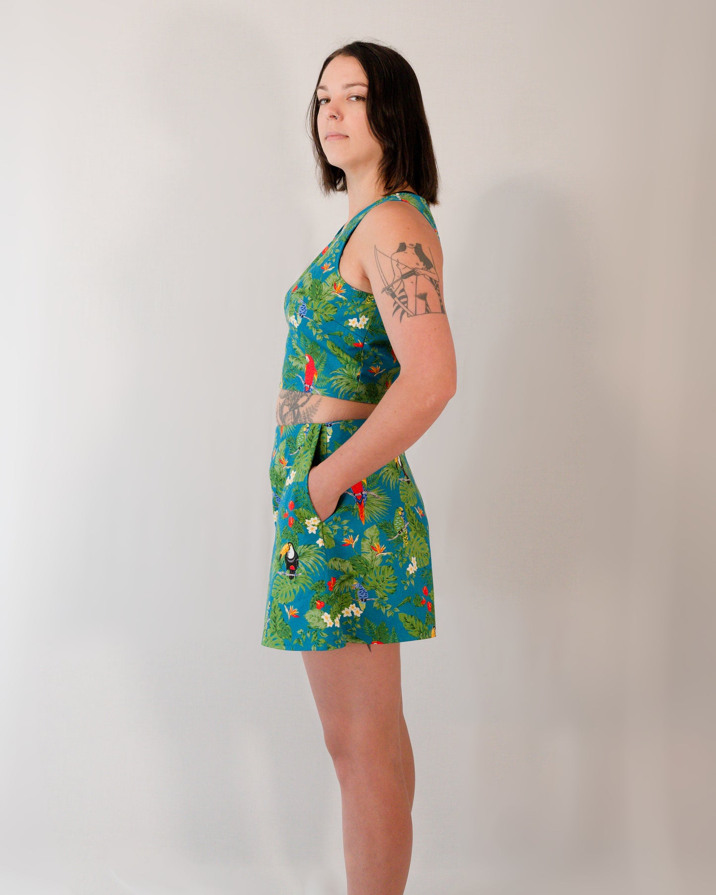 Left side of flared shorts in a turquoise parrot print. Model has hands in pockets. Paired with matching sleeveless crop top.