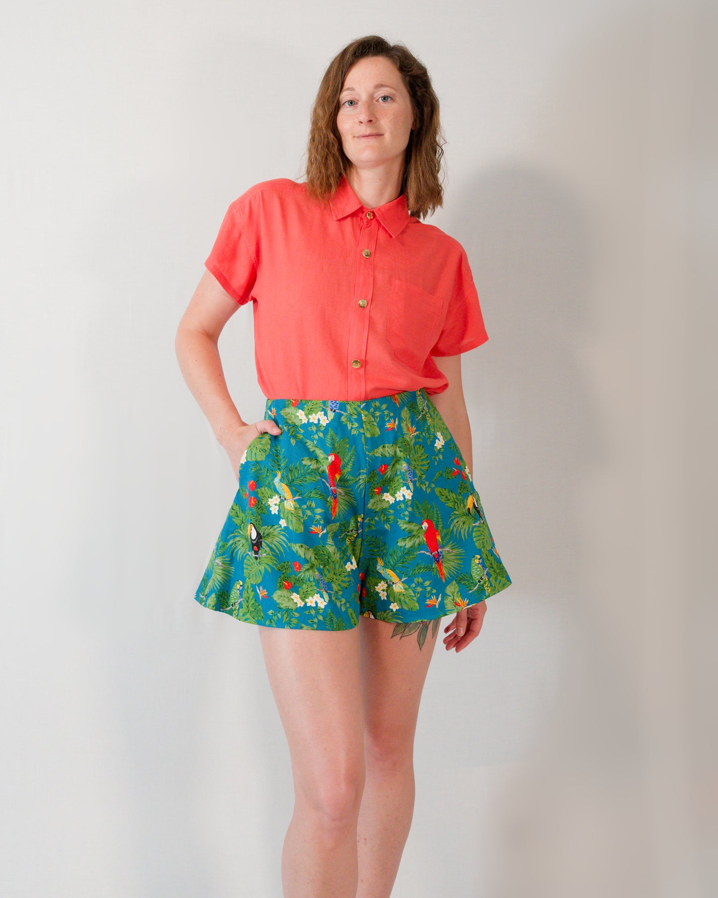 Front of flared shorts in a turquoise parrot print. Paired with a short-sleeved blouse in callistemon red, tucked in.