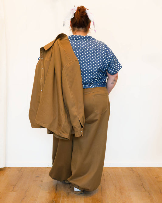 Hannah, facing backwards against a white wall, modelling khaki wide-leg jeans, a blue polkadot shirt and holding a khaki chore jacket over her shoulder.