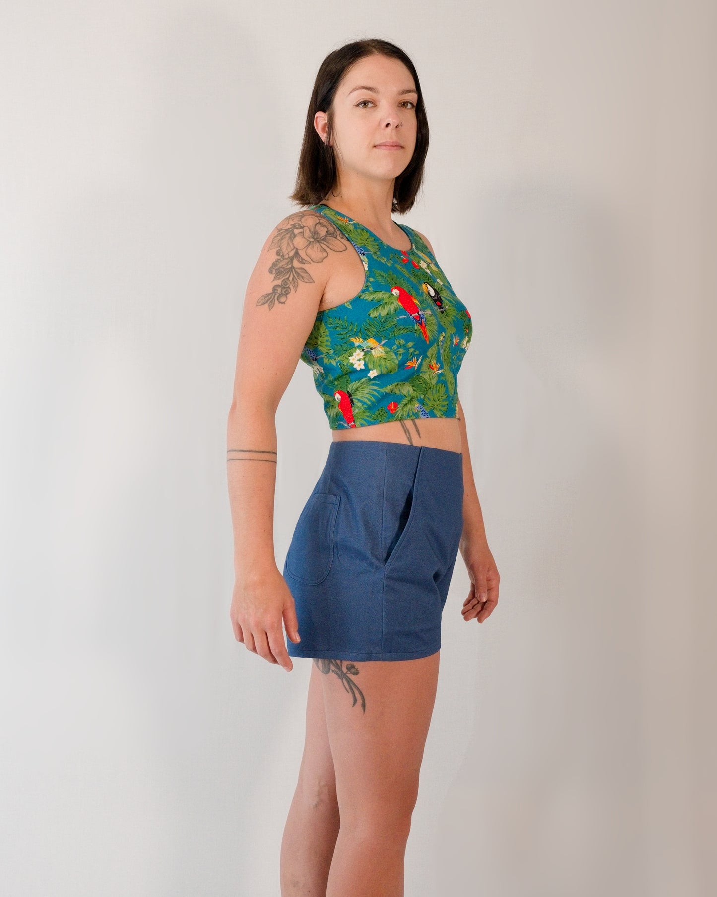 Right side of Kingfisher A-line shorts in dark blue. Visible front and back pockets. Paired with sleeveless crop top in turquoise parrot print.