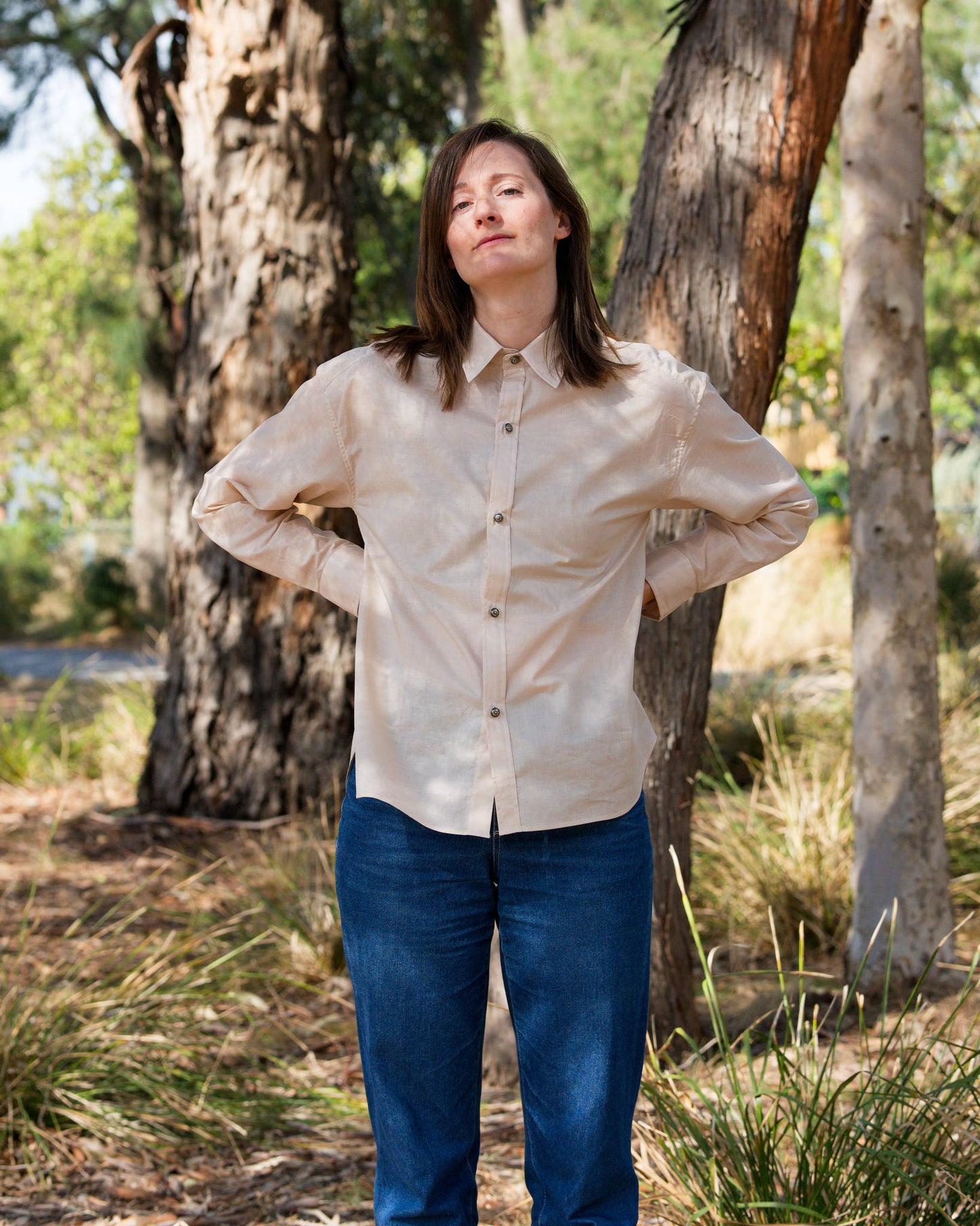 Rachael modelling a champagne-coloured button-down shirt and blue jeans in an outdoor setting with trees and long grass.