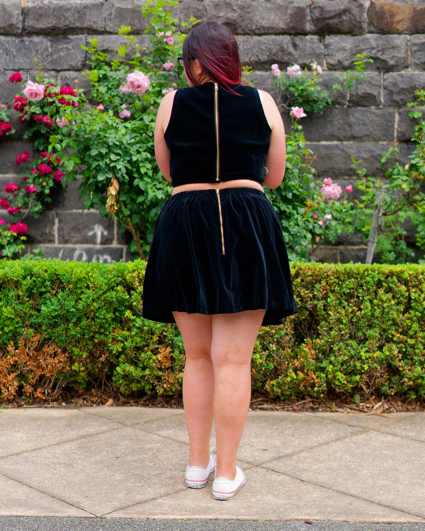 Veronica modelling a black velvet sleeveless crop top and mini skirt with white sneakers, showing golden zippers on the back and facing a bluestone wall and rose bushes.