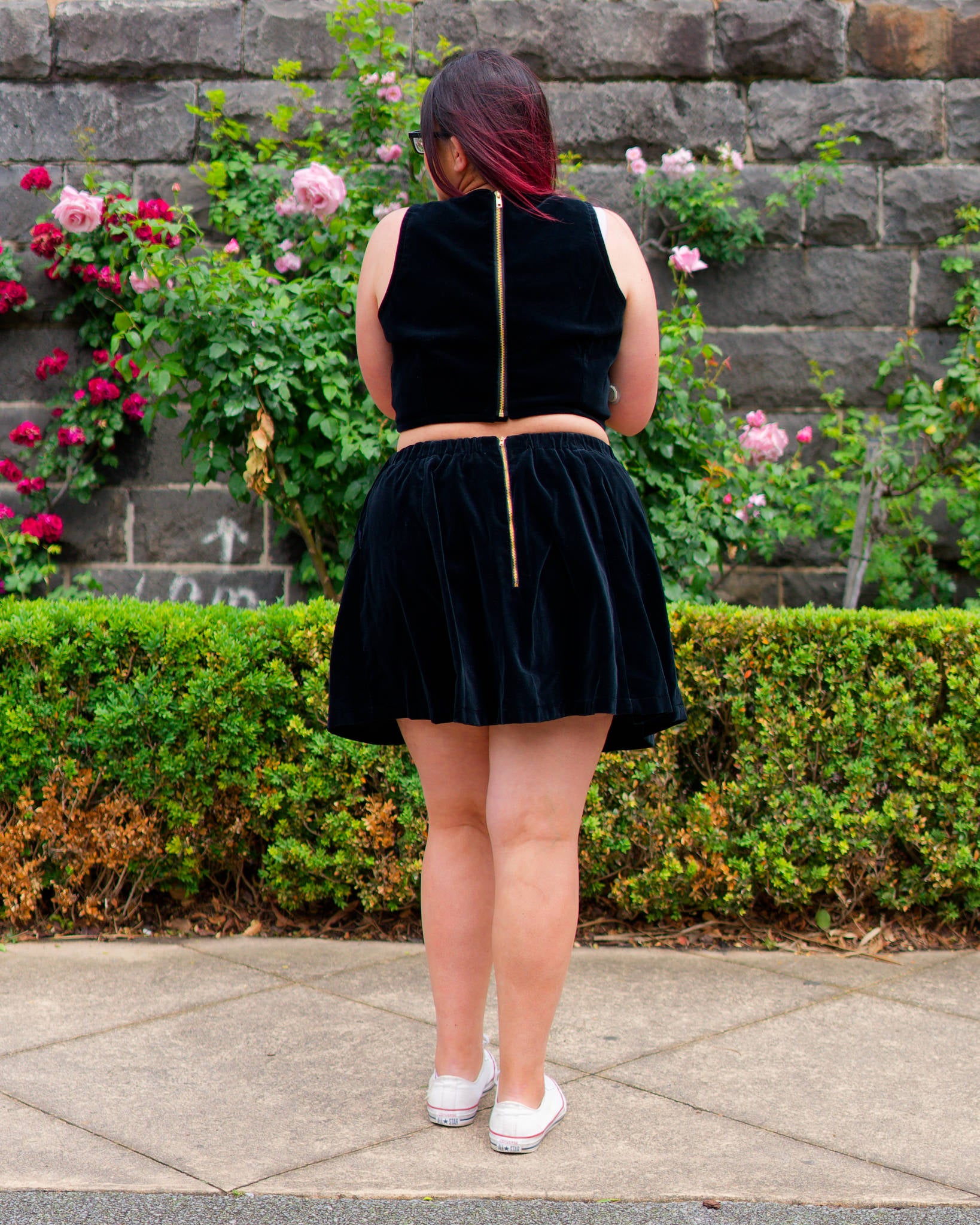 Veronica modelling a black velvet sleeveless crop top and mini skirt with white sneakers, showing golden zippers on the back and facing a bluestone wall and rose bushes.