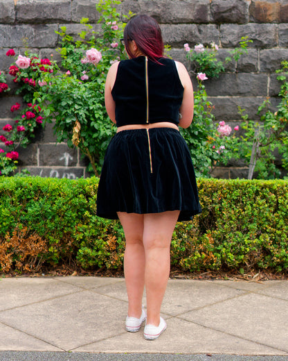 Veronica modelling a black velvet sleeveless crop top and mini skirt with white sneakers, showing golden zippers on the back and facing a bluestone wall and rose bushes.