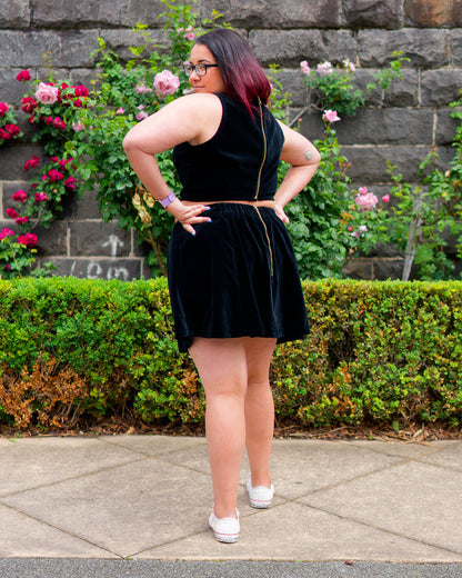 Veronica, looking backwards, modelling a black velvet sleeveless crop top and mini skirt with white sneakers, standing in front of a bluestone wall and rose bushes.