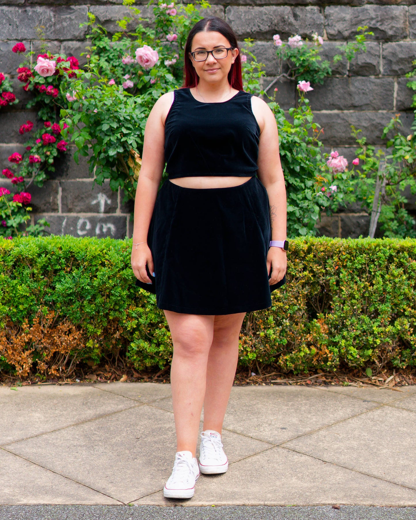 Veronica modelling a black velvet sleeveless crop top and mini skirt with white sneakers, standing in front of a bluestone wall and rose bushes.
