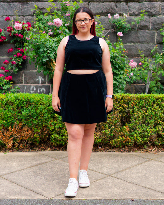 Veronica modelling a black velvet sleeveless crop top and mini skirt with white sneakers, standing in front of a bluestone wall and rose bushes.