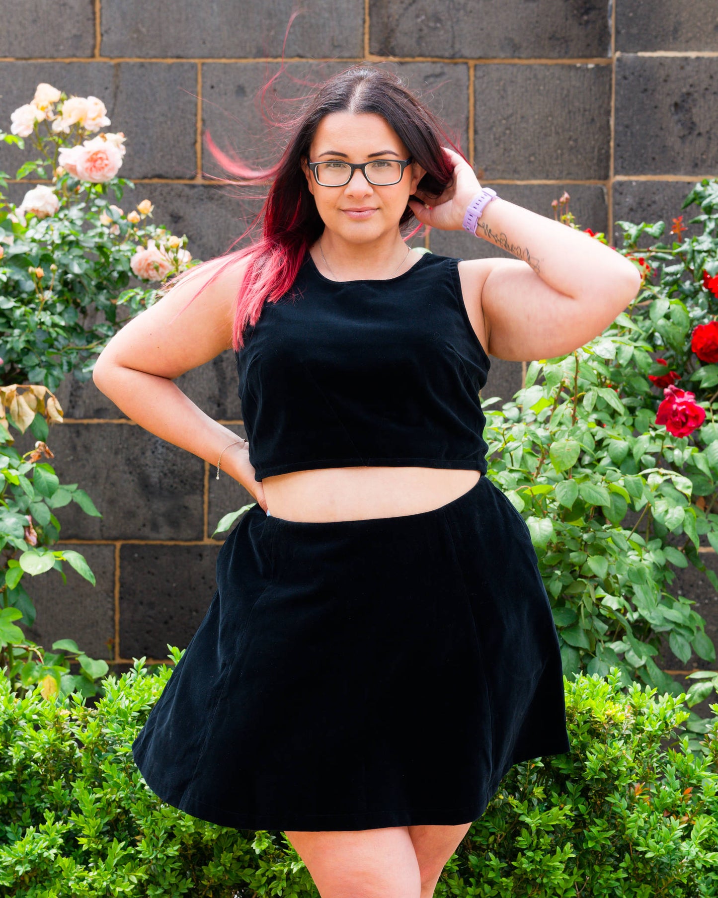 Veronica modelling a black velvet sleeveless crop top and mini skirt, standing in front of a bluestone wall and rose bushes.