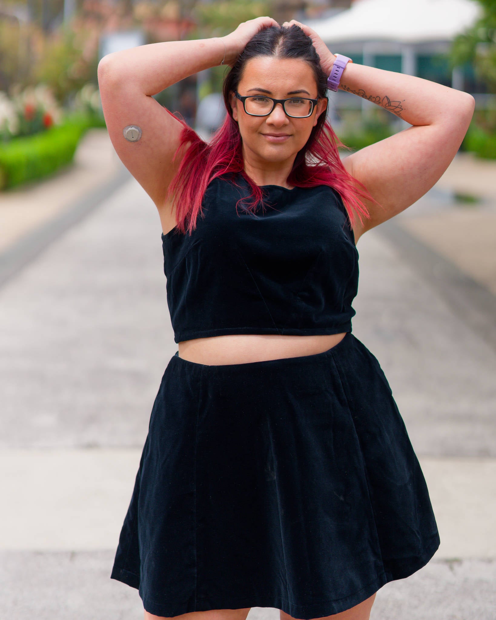 Veronica with hands behind her head, standing in a leafy promenade, modelling a black velvet sleeveless crop top and mini skirt