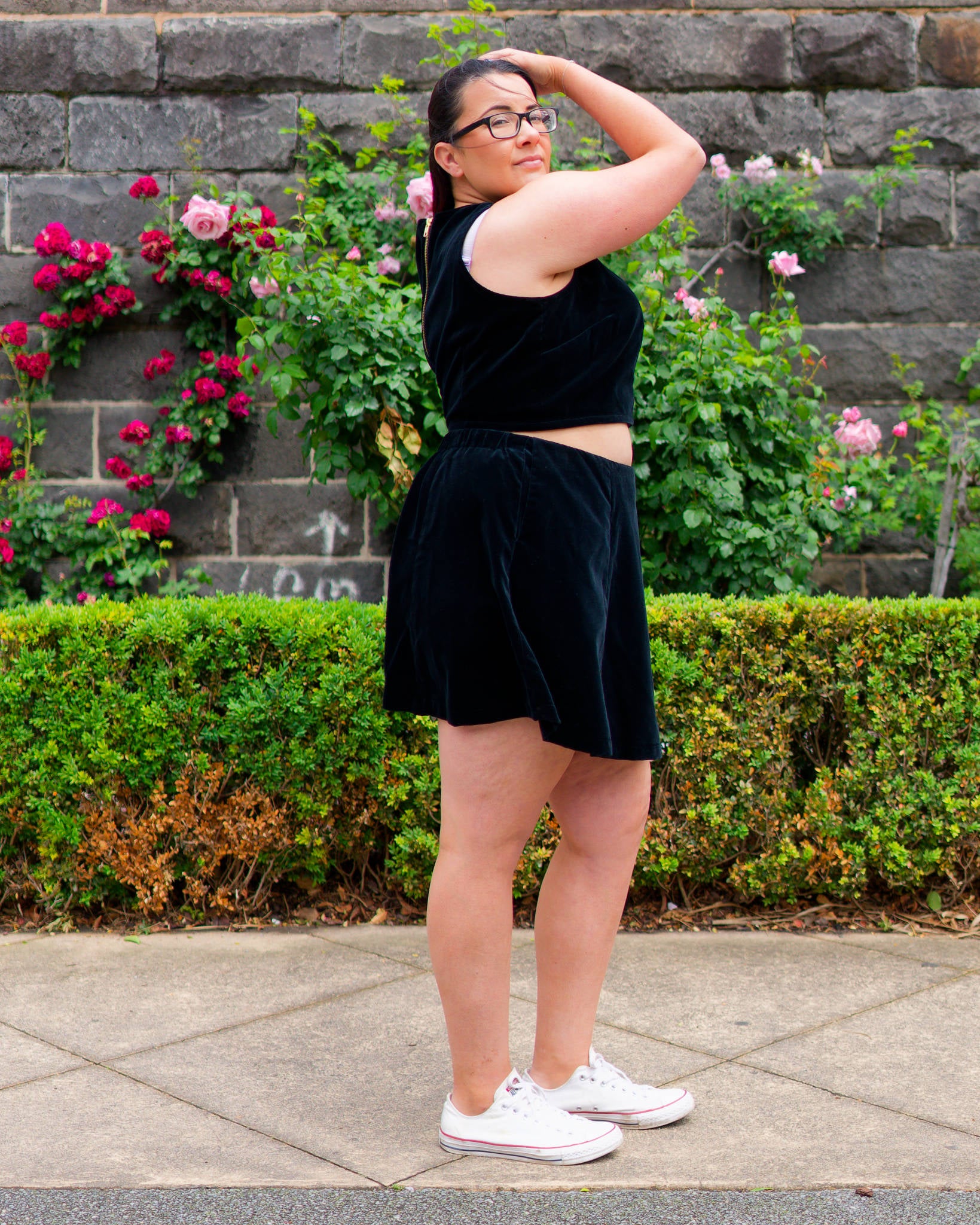 Veronica, standing side-on, modelling a black velvet sleeveless crop top and mini skirt with white sneakers, standing in front of a bluestone wall and rose bushes.