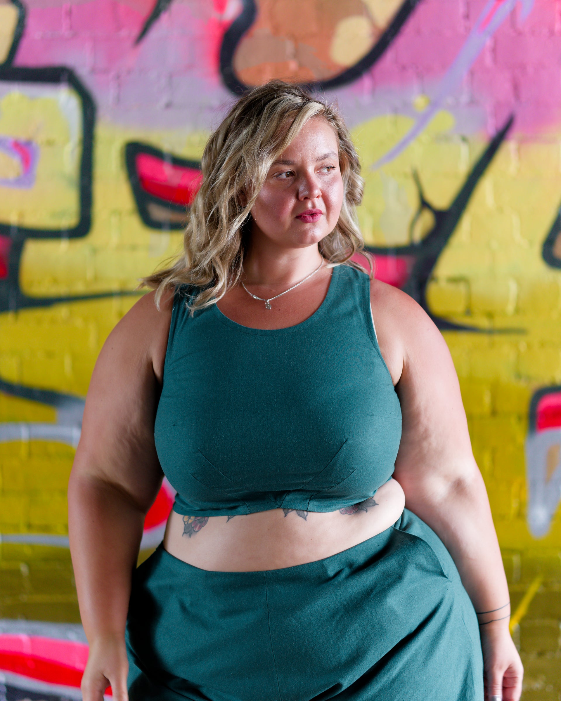 Front of a sleeveless crop top in deep emerald. Two waist darts and a bust dart for shaping. Graffiti wall in background.