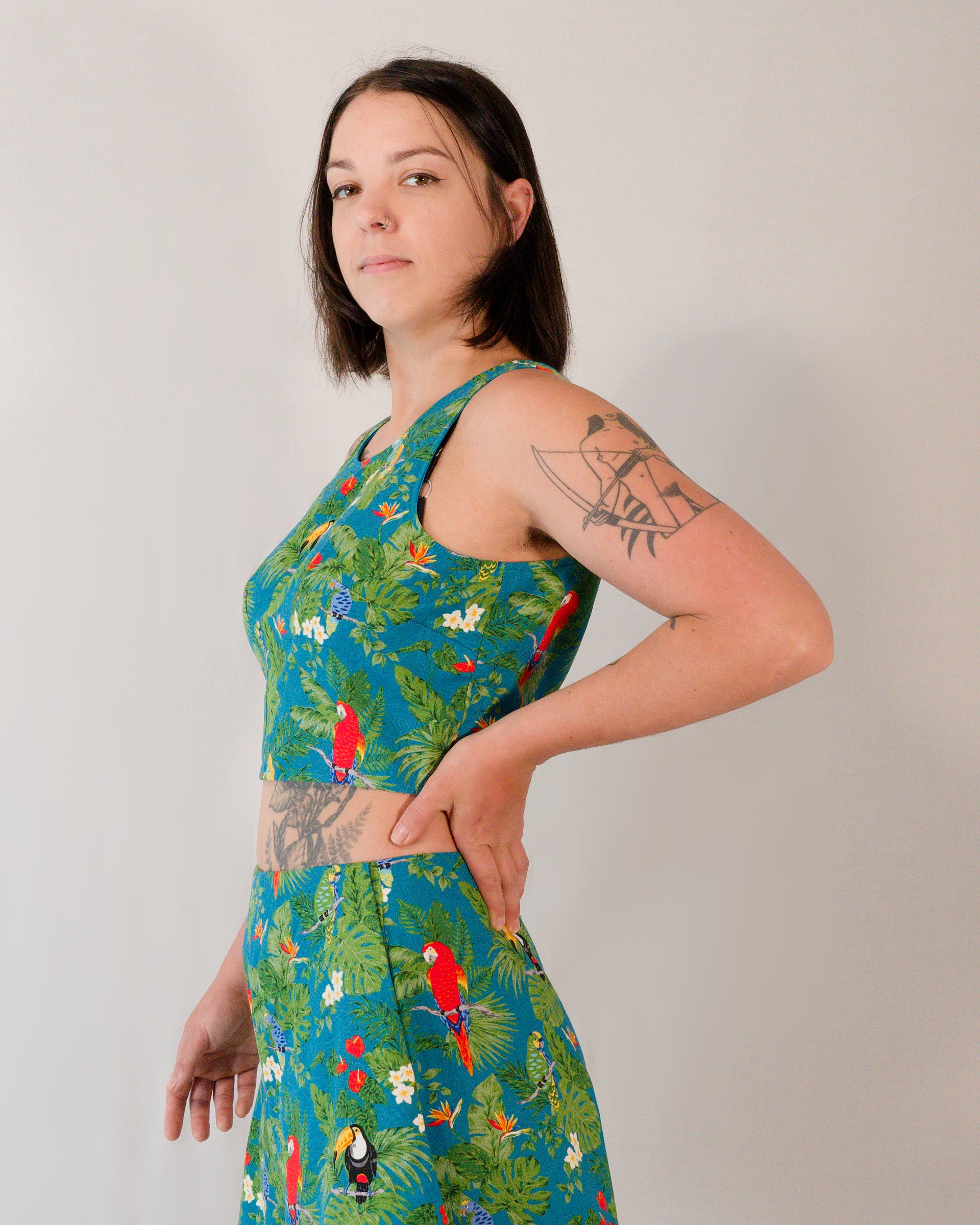 Left side of a sleeveless crop top in a turquoise print with plants, parrots and toucans.