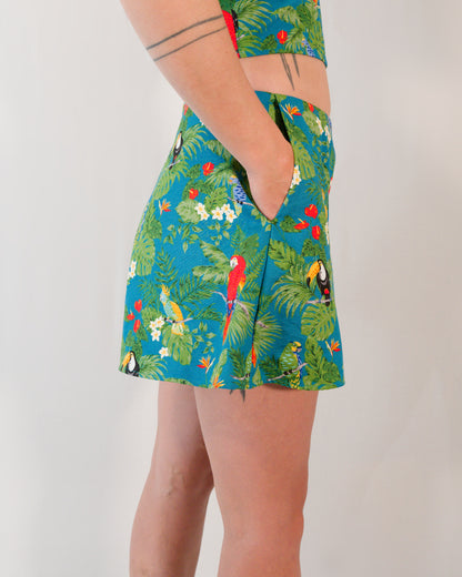 Right side of flared shorts in a turquoise print with plants, parrots and toucans. Model has hand in pocket.