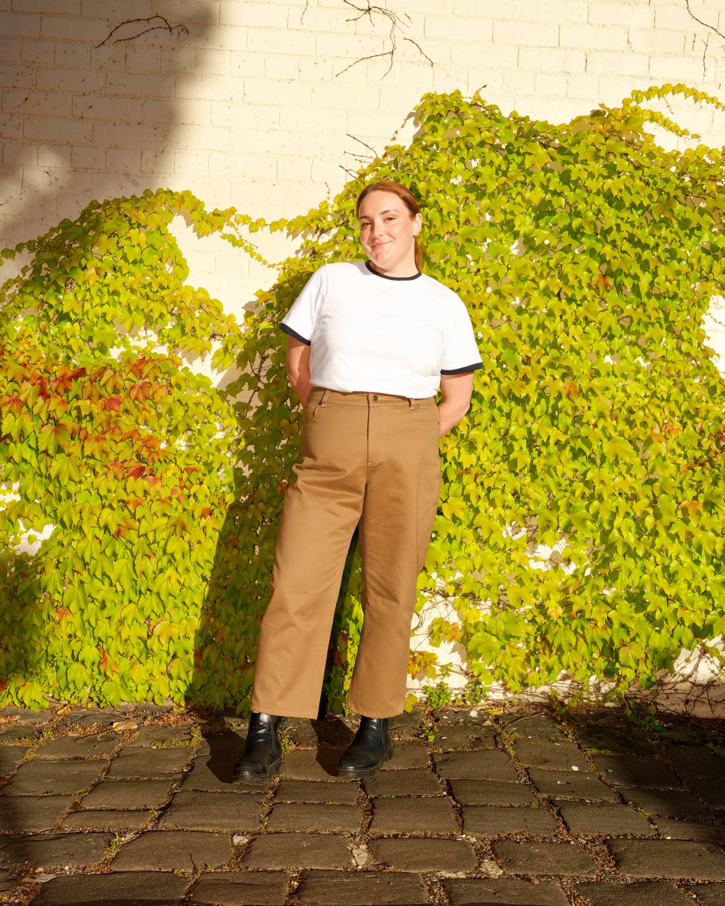 Lydia, modelling a white t-shirt with navy trim tucked into khaki barrel-leg jeans, standing in front of a green ivy wall.