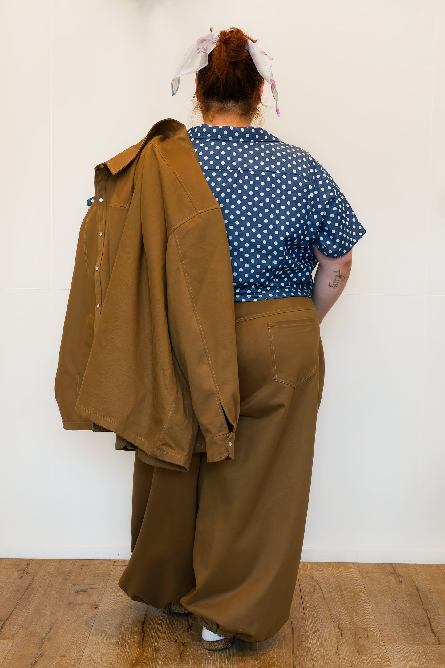 Hannah, facing backwards against a white wall, modelling khaki wide-leg jeans, a blue polkadot shirt and holding a khaki chore jacket over her shoulder.