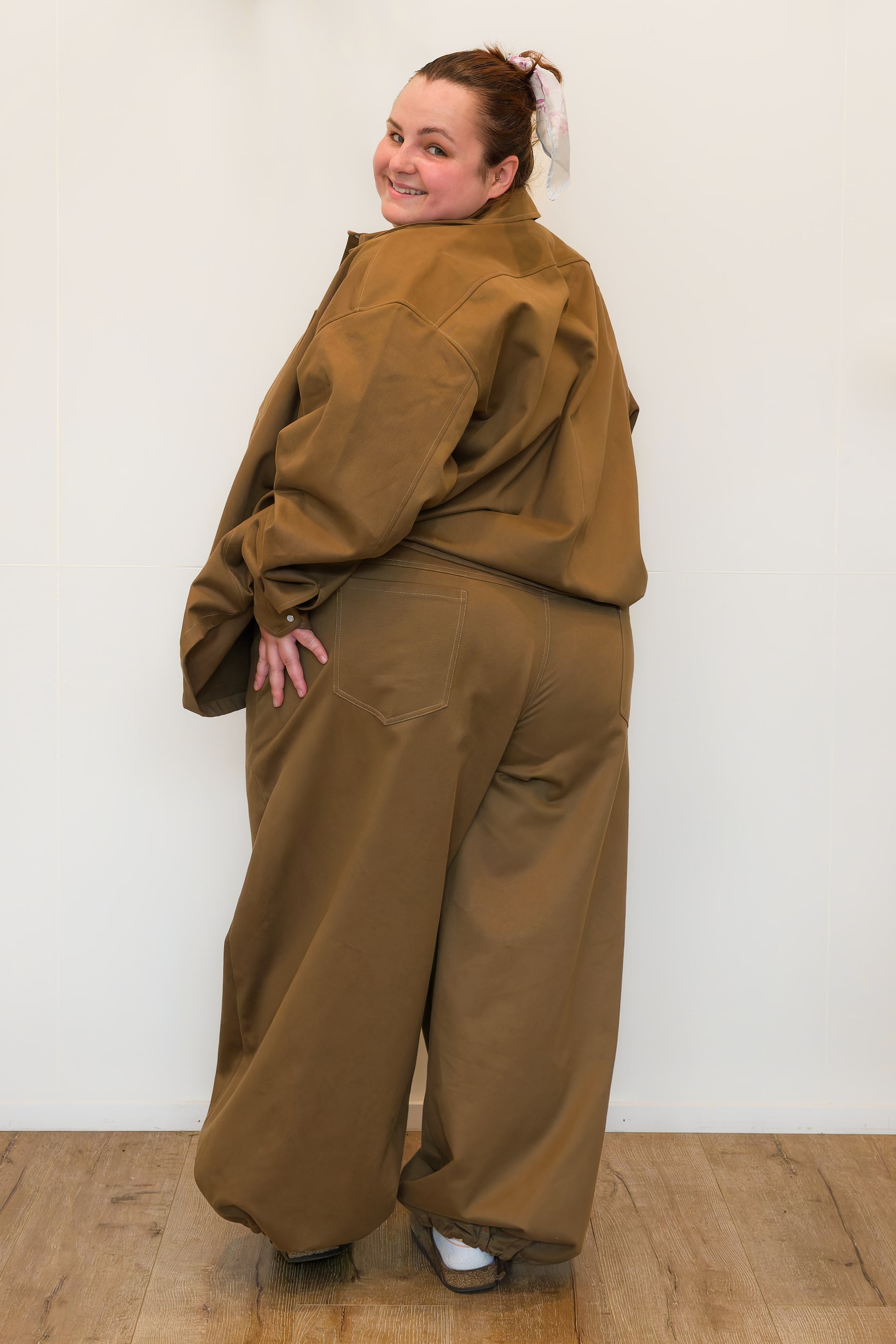 Hannah, facing backwards against a white wall, modelling khaki wide-leg jeans and a khaki chore jacket.