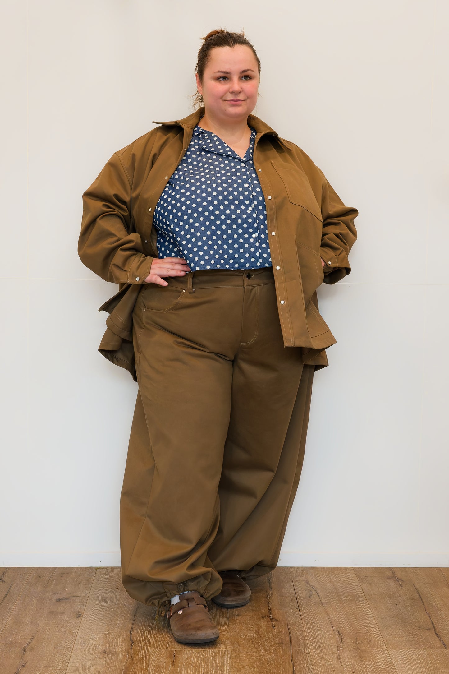 Hannah, in front of a white wall, modelling khaki wide-leg jeans and a khaki chore jacket over a a blue polkadot shirt.