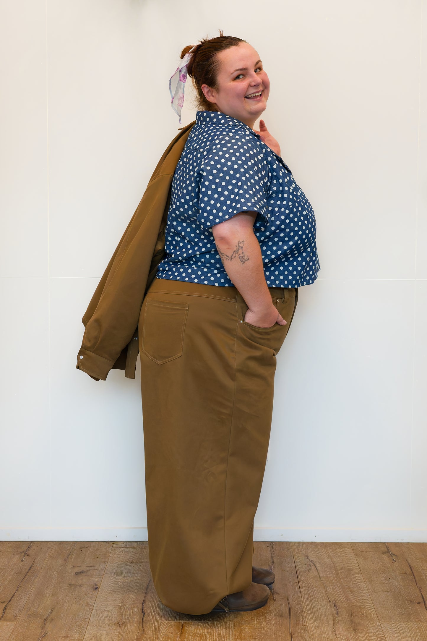 Hannah, side-on in front of a white wall, modelling khaki wide-leg jeans, a blue polkadot shirt and holding a khaki chore jacket over her shoulder.
