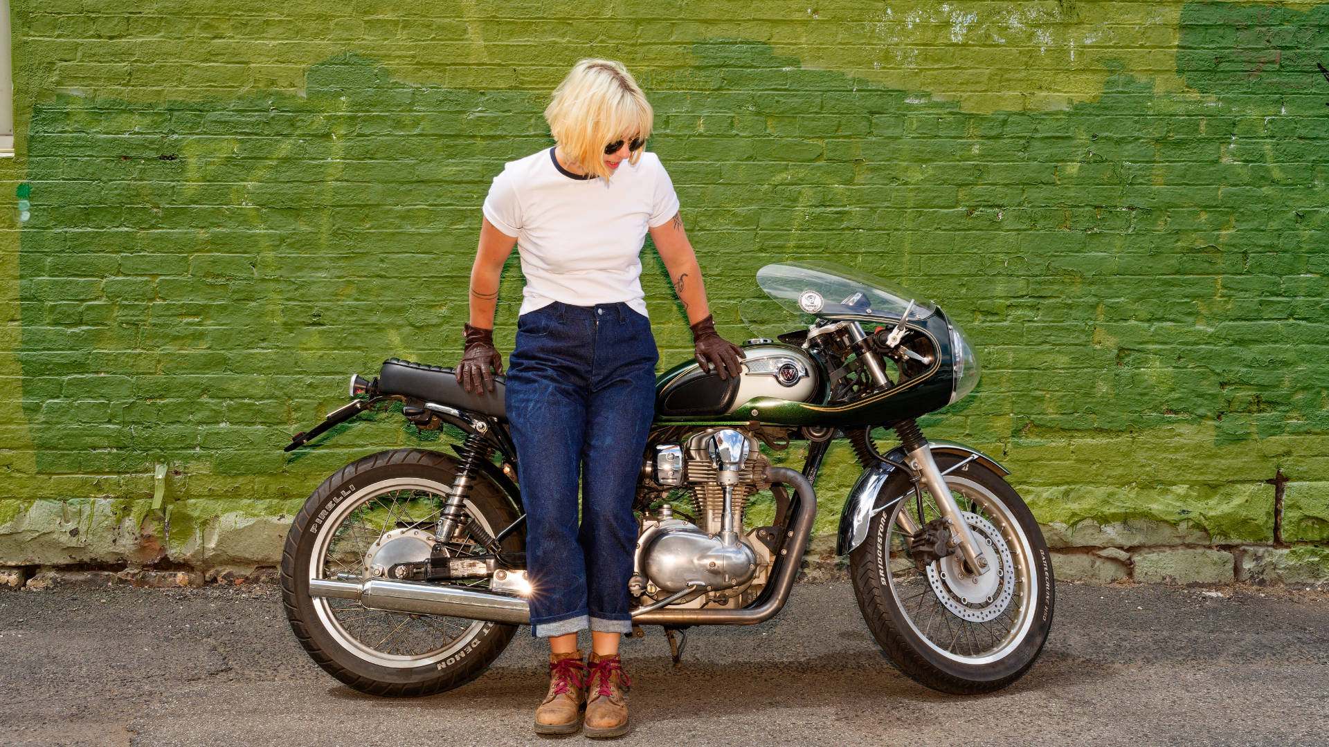 Alex, modelling a white t-shirt and blue jeans, standing next to a vintage motorcycle against a green brick wall.
