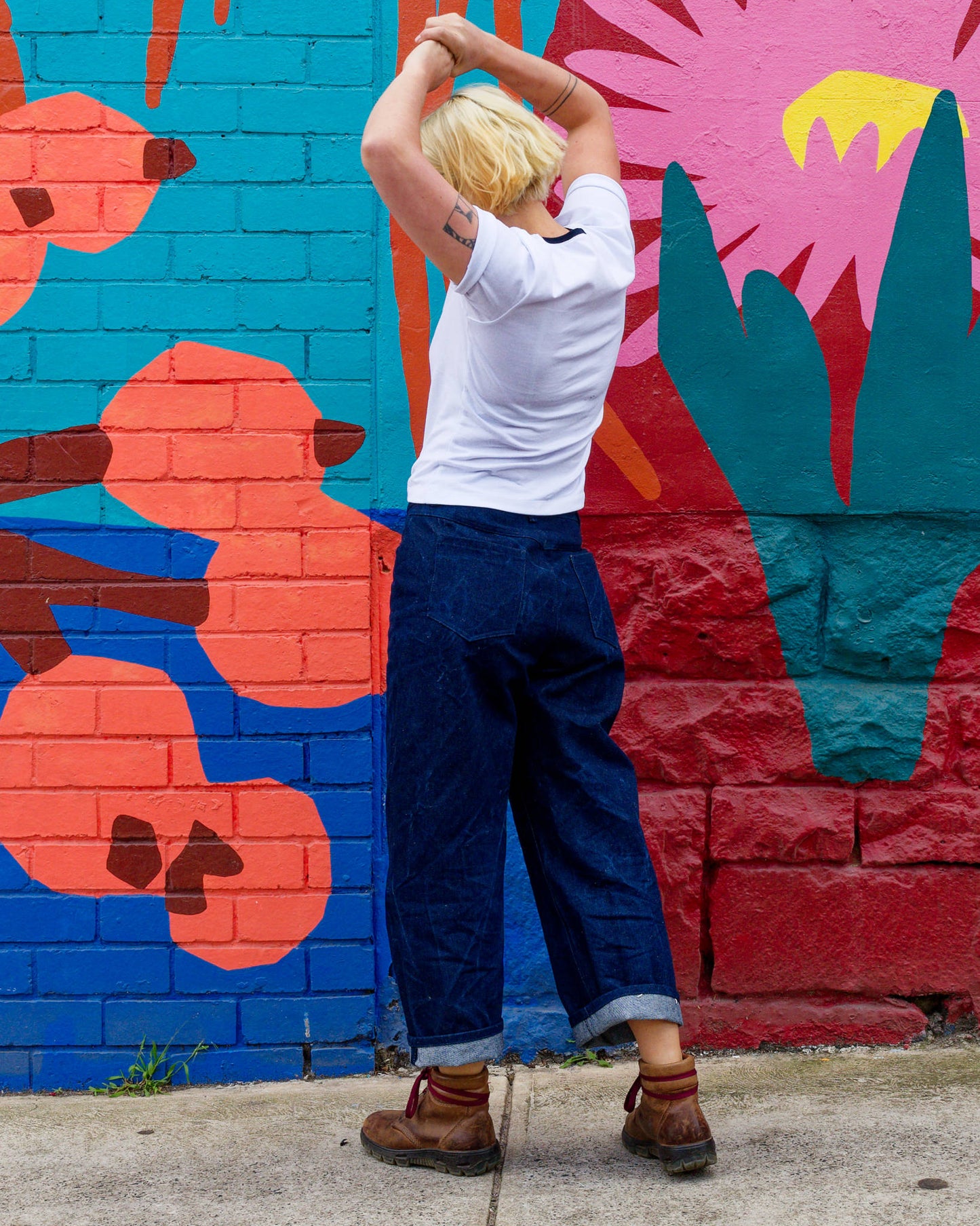 Alex modelling a white t-shirt, blue jeans, and brown boots, holding her hands above her head while standing in front of a colourful mural.