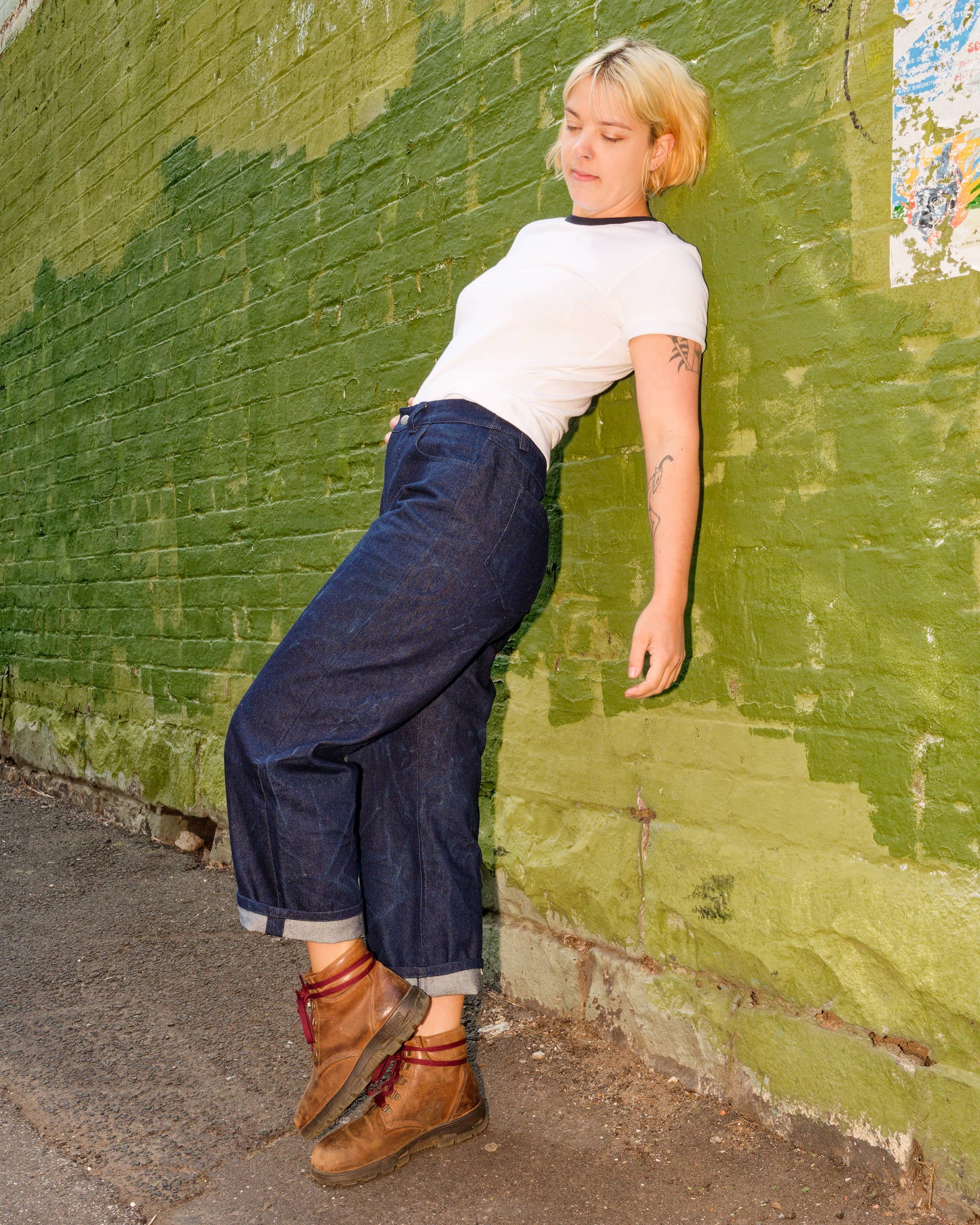 Alex modelling a white t-shirt, blue jeans, and brown boots and leaning against a green brick wall.