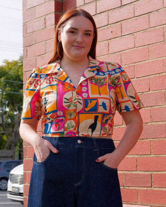 Woman wearing a colorful patterned shirt standing against a brick wall.